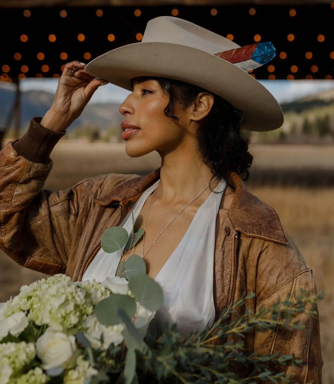 A bride in a cowboy hat and leather jacket under the hangar string lights