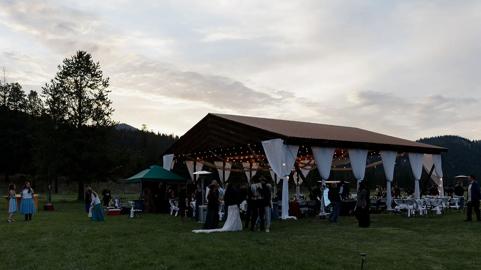 Wedding guests gathering at the open-air hangar at dusk, white drapes and string lights inside